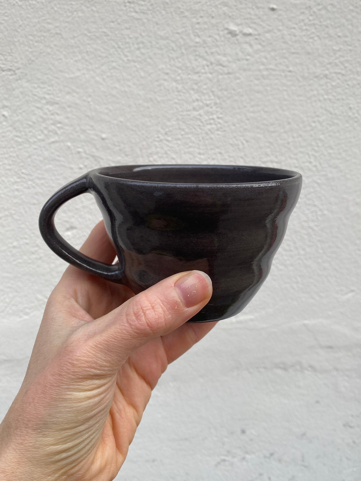 Hand holding a black ceramic cup with soft shapes and a handle against a light gray background