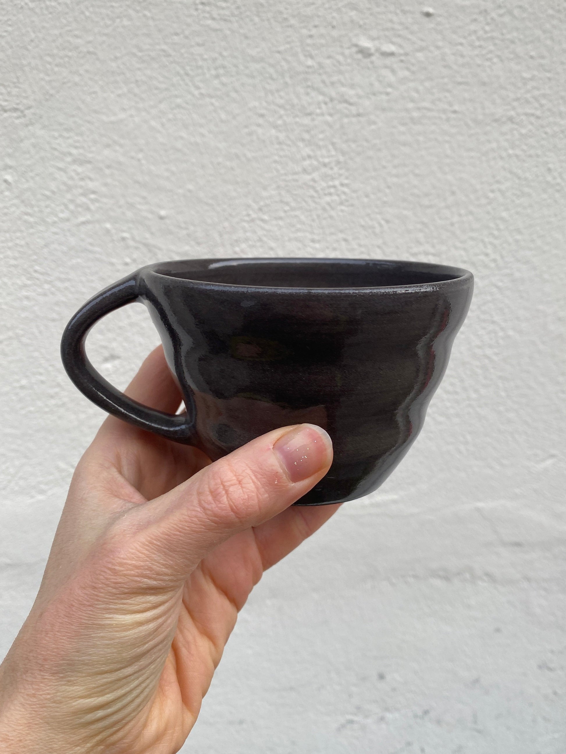 Hand holding a black ceramic cup with soft shapes and a handle against a light gray background