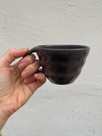 Hand holding a dark grey ceramic cup against a white wall