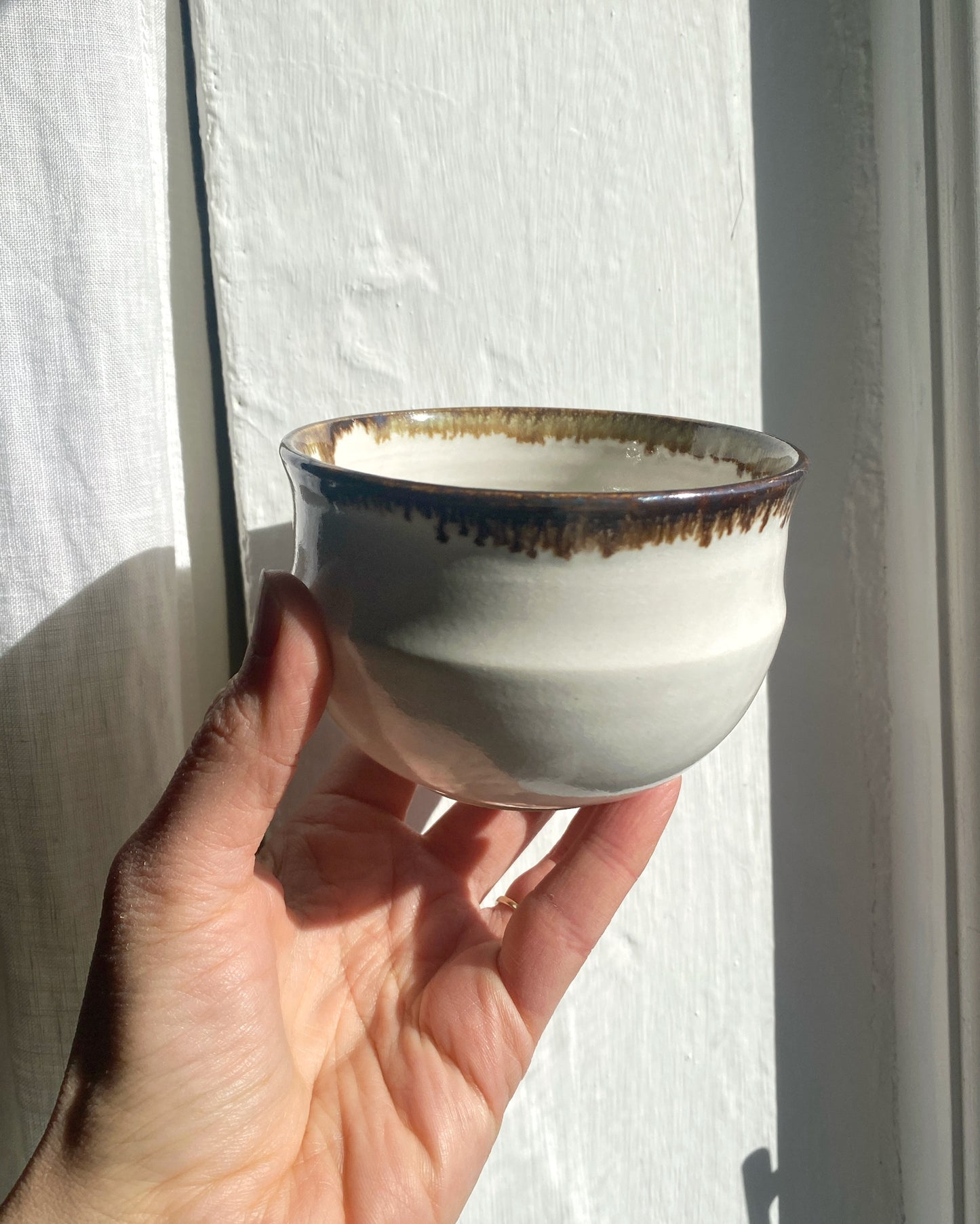 A hand holding a small handmade ceramic bowl with a matte off-white body and a dark brown glazed drip rim, set against a white textured wall with natural sunlight and shadows.