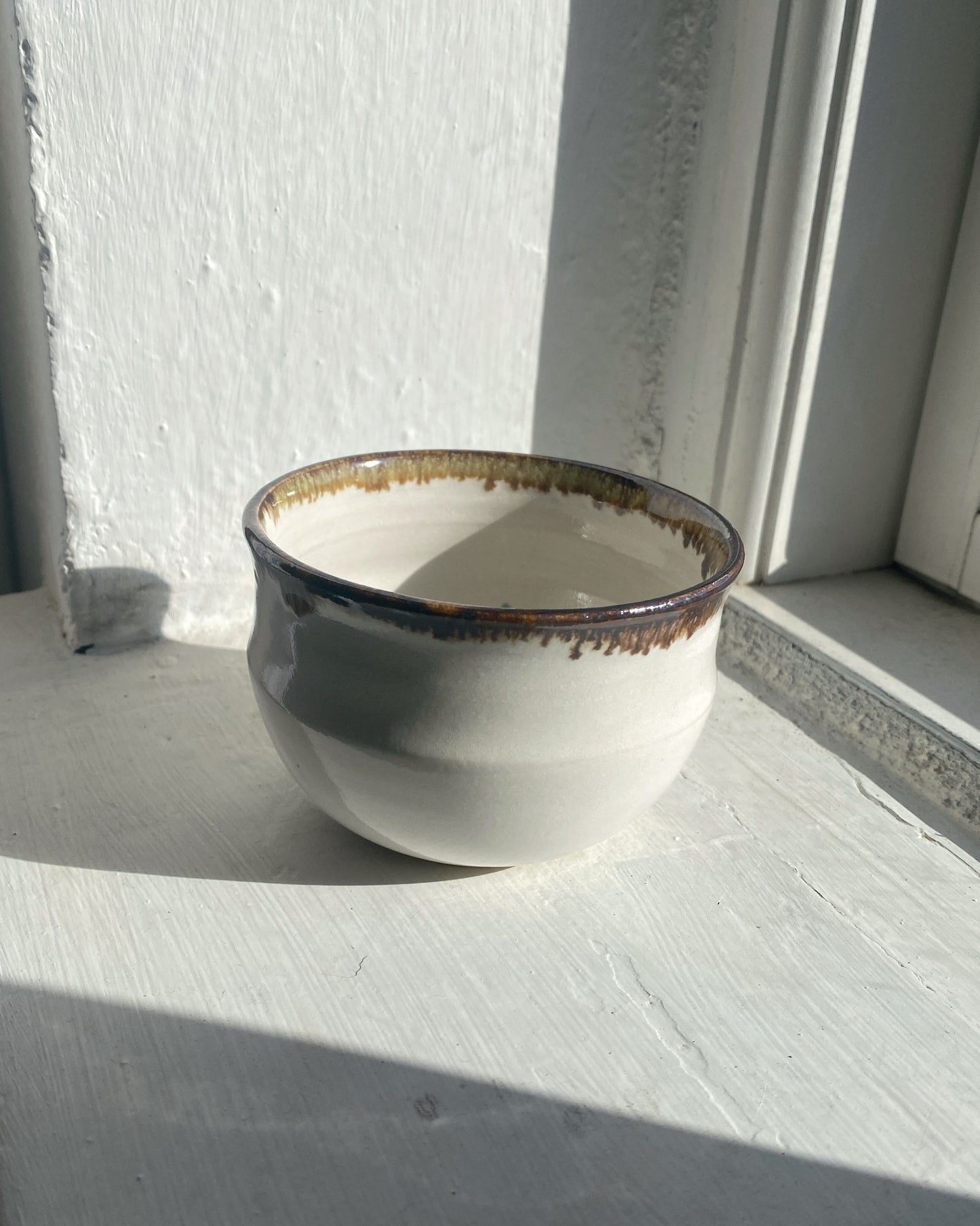 A handmade ceramic cup with a rounded shape, featuring a glossy off-white glaze, a dark brown rim with gold-toned speckling, and a rustic dripping edge, set on a white painted wooden surface next to a window.