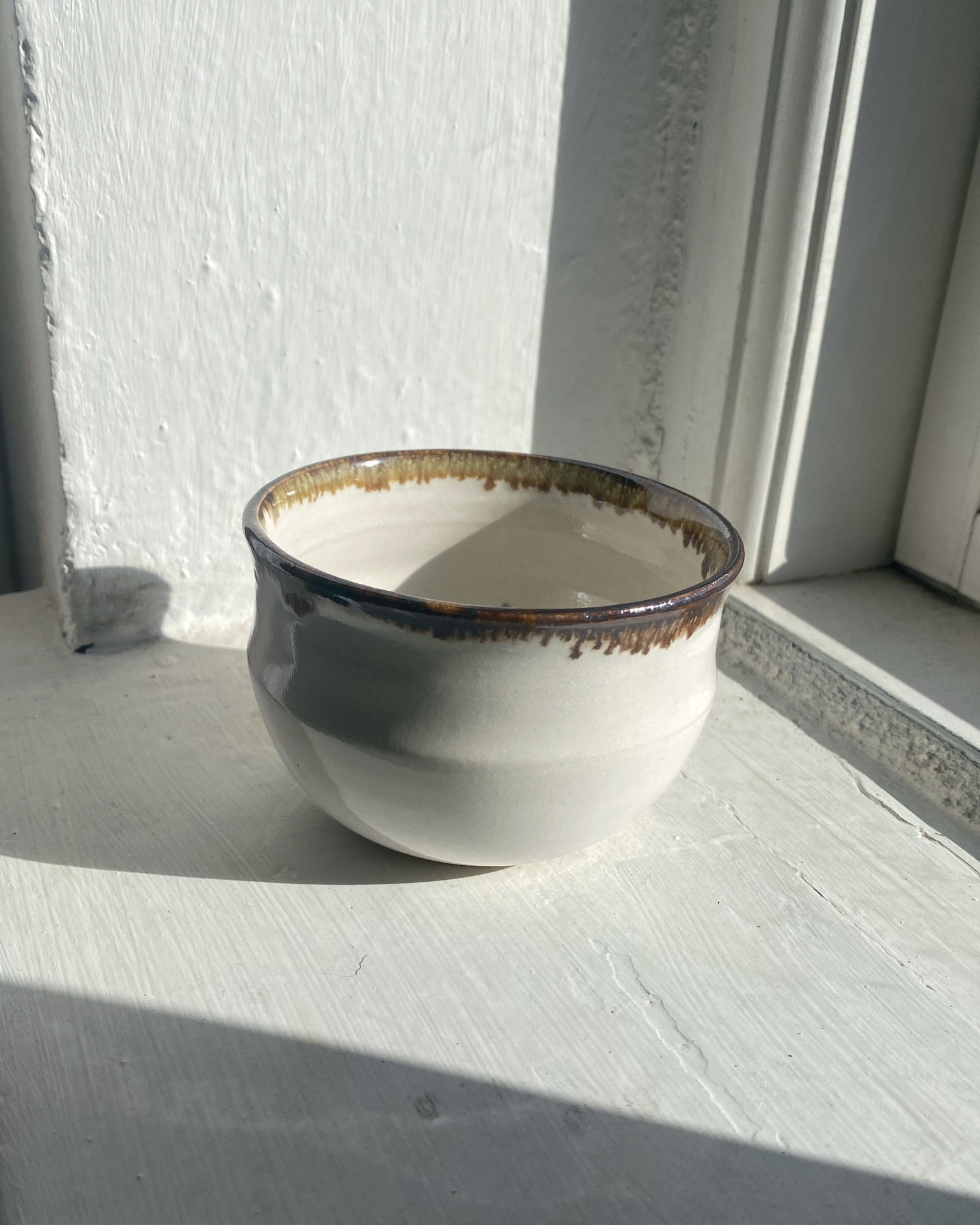 A handmade ceramic cup with a rounded shape, featuring a glossy off-white glaze, a dark brown rim with gold-toned speckling, and a rustic dripping edge, set on a white painted wooden surface next to a window.
