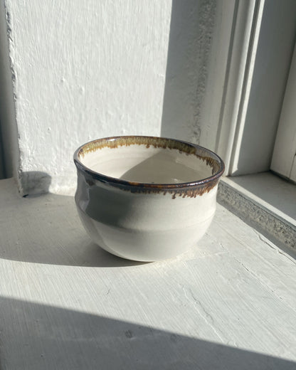 A handmade ceramic cup with a rounded shape, featuring a glossy off-white glaze, a dark brown rim with gold-toned speckling, and a rustic dripping edge, set on a white painted wooden surface next to a window.